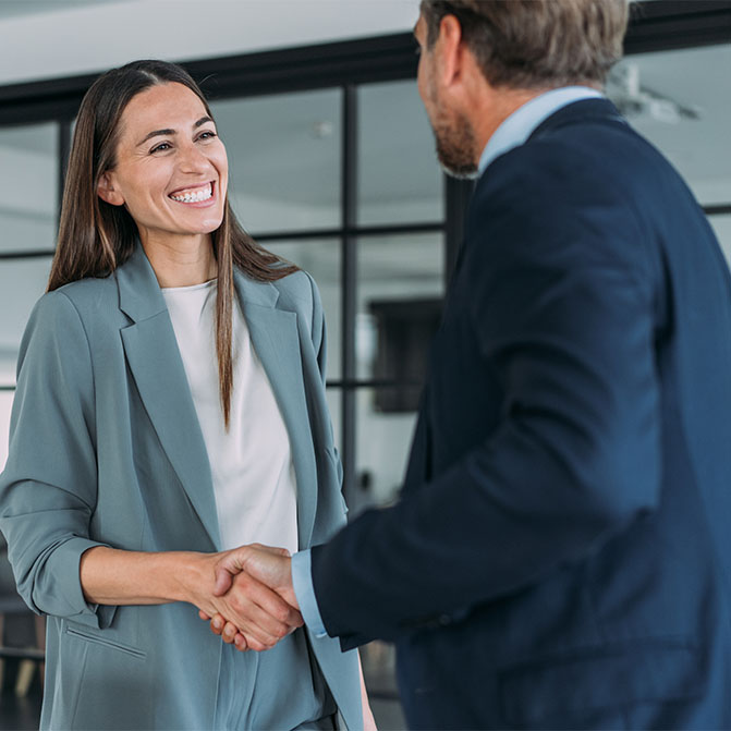 Zwei Geschäftsleute – eine brünette Frau, die uns zugewandt ist, und ein Mann, der uns den Rücken zugewandt hat – schütteln sich die Hände.